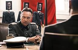 En man sitter vid ett skrivbord i uniform. Bakom sitter fotografier på personer på pinnar.